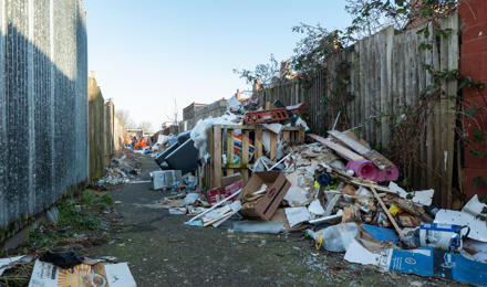 Narrow Alley Littered With Scattered Garbage, Broken Items, And Cardboard Boxes Along Wooden Fences