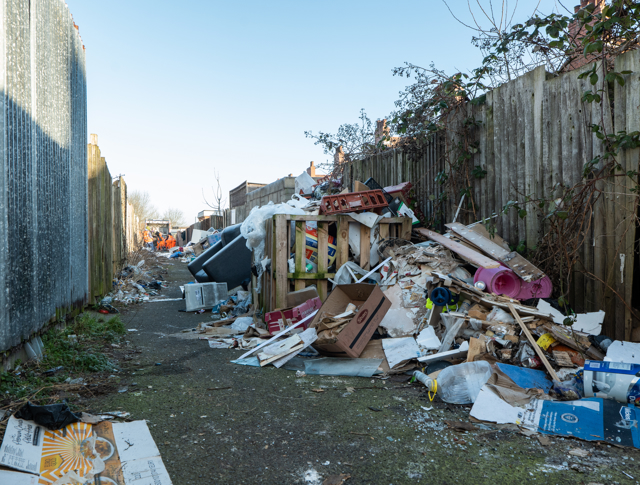 Narrow Alley Littered With Scattered Garbage, Broken Items, And Cardboard Boxes Along Wooden Fences