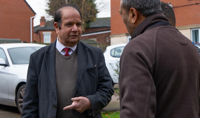 Two Men Stood In A Car Park Talking With One Facing The Camera And With His Back To The Camera