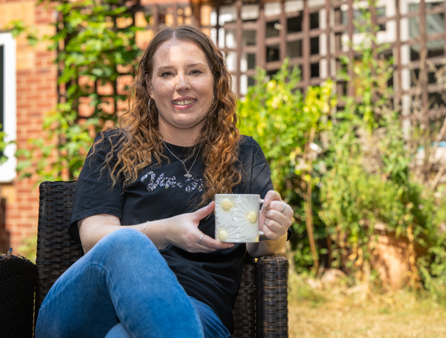 Woman With Curly Hair Smiling, Seated In A Wicker Chair Outdoors. She Holds A Mug With A Daisy Design. Garden And Brick Wall In The Background