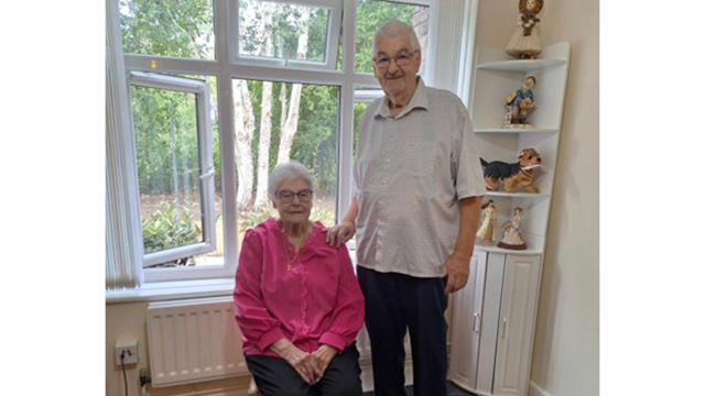 Older Couple Posing For Camera With Man Standing Anf Woman Sat On A Chair