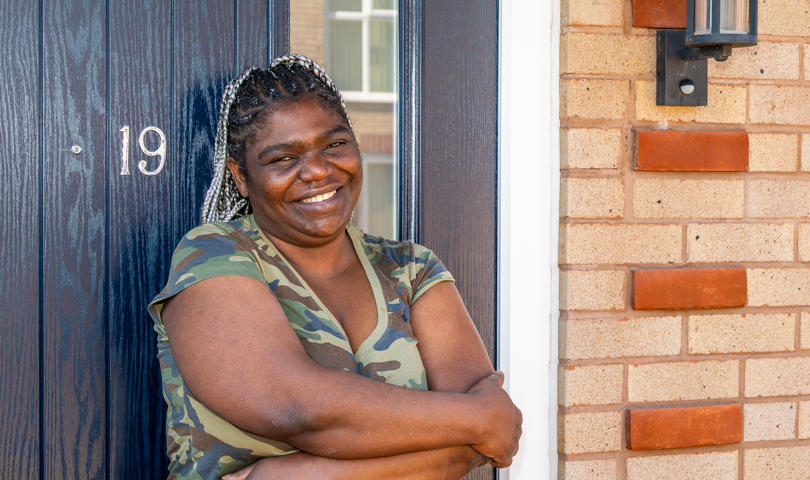 A Person Stands With Arms Folded In The Doorway Of A Brick House, Next To A Dark Blue Front Door Numbered 19, With An Outdoor Wall Light Mounted Beside The Door