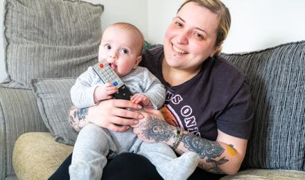 A Person Is Sitting On A Sofa, Holding A Baby Who Is Chewing On A Remote Control. Both Are Smiling. The Person Has Tattoos On Their Arms