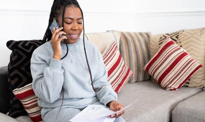 A Person With Long Braided Hair Is Sitting On A Sofa, Holding A Phone To Their Ear. They Are Dressed In A Grey Tracksuit And Holding Papers