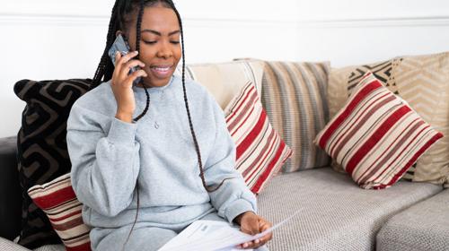 A Person With Long Braided Hair Is Sitting On A Sofa, Holding A Phone To Their Ear. They Are Dressed In A Grey Tracksuit And Holding Papers