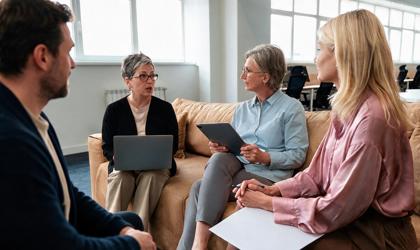 Three Women One Man Sat On Sofa With Laptops Having A Meeting In An Office Setting