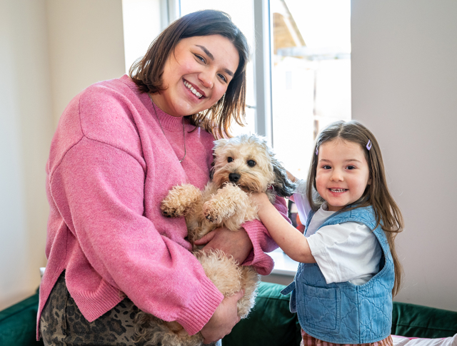 Two Smiling Individuals And A Small Dog Indoors, With One Person Holding The Dog. Both Are Looking At The Camera