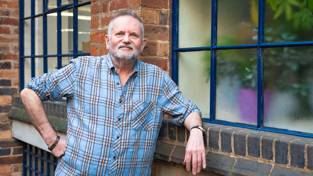 Tenant Wearing A Blue And White Checkered Shirt Leaning Against A Window Ledge