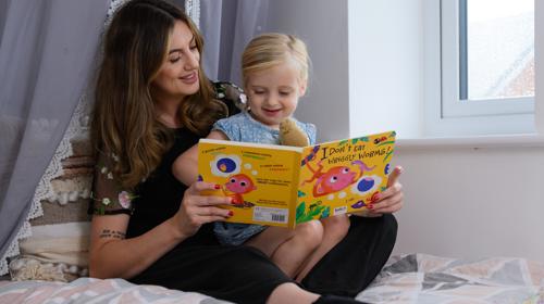 Mother And Daughter Sitting On A Bed Reading A Book Called I Don't Eat Wriggly Worms
