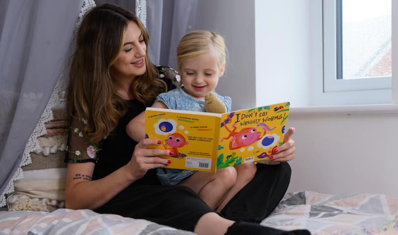 Mother And Daughter Sitting On A Bed Reading A Book Called I Don't Eat Wriggly Worms