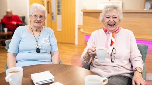 Two Elderly Individuals Smiling And Holding Mugs At A Table In A Bright Room, With Other People Visible In The Background