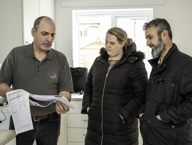 A Man In A Dark Polo Shirt Shows Paperwork To A Man And Woman Wearing Dark Winter Coats In A Kitchen